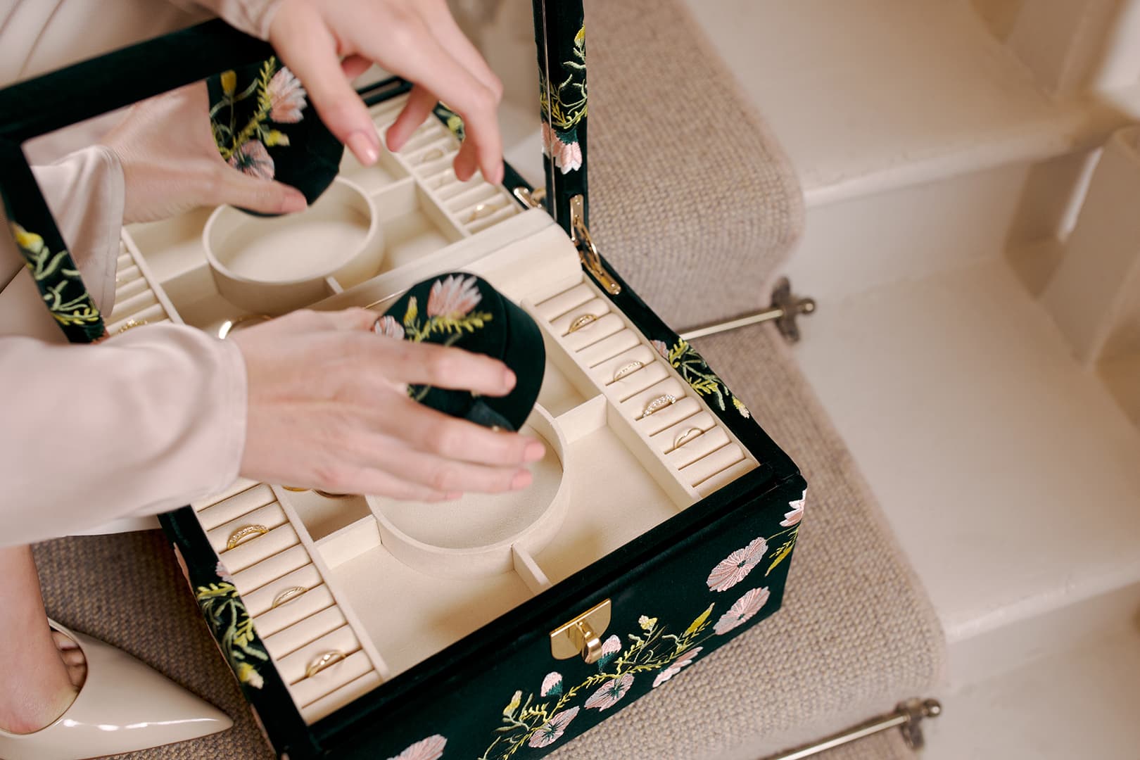 WOLF Zoe Jewellery Box lifestyle image. Hands arranging jewellery inside an open WOLF Zoe Jewellery Box with floral embroidered exterior and organised storage compartments.